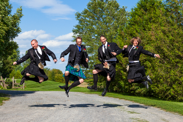 Four groomsmen wearing kilts jump in the air with excitement