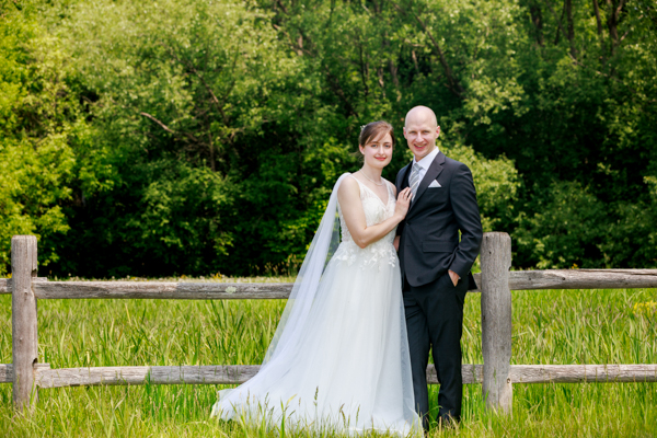 A bride and groom pose in a feild next to a wood rail frence
